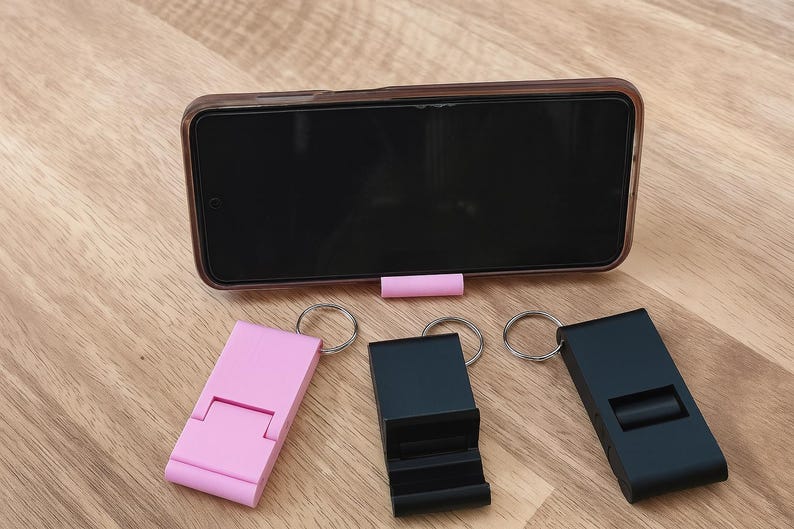 K&ouml;nnte beinhalten: Ein Smartphone liegt auf einem rosa St&auml;nder, mit drei Schl&uuml;sselanh&auml;ngern in Rosa und Schwarz. Die Schl&uuml;sselanh&auml;nger sind als Telefonst&auml;nder konzipiert. Die Artikel befinden sich auf einer Holzoberfl&auml;che.
