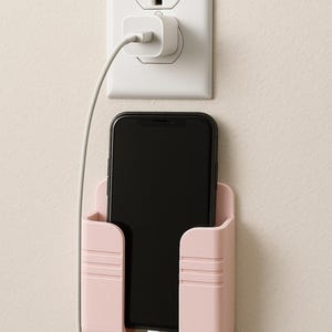 May include: A pink phone holder with a black smartphone inside, plugged into a white charger and outlet. The holder is mounted on a beige wall, providing a convenient charging station.
