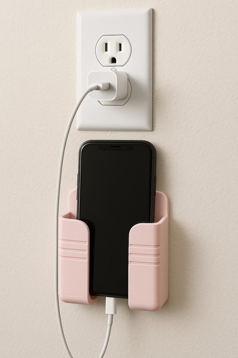 May include: A pink phone holder with a black smartphone inside, plugged into a white charger and outlet. The holder is mounted on a beige wall, providing a convenient charging station.