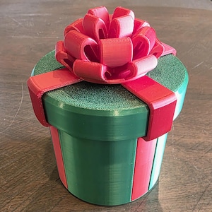 Puede incluir: Una caja de regalo cilíndrica verde con una cinta y un lazo rojos. La caja tiene una parte superior texturizada y rayas rojas verticales. El lazo es grande y detallado, con múltiples bucles. La caja de regalo es una decoración festiva.