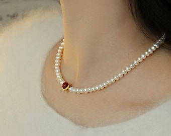May include: A delicate pearl necklace with a small, round, red gemstone set in a gold-colored metal. The necklace is composed of numerous small, white pearls. The necklace is worn on a person's neck.