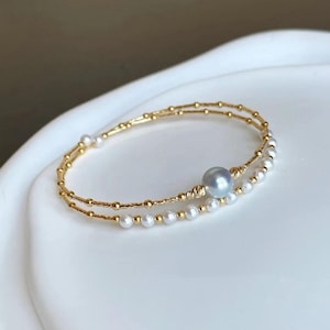 May include: A delicate gold bracelet with two parallel strands. One strand is adorned with small, round pearls, while the other features tiny gold beads. A larger, iridescent gray pearl is centered on the bracelet, adding a focal point.