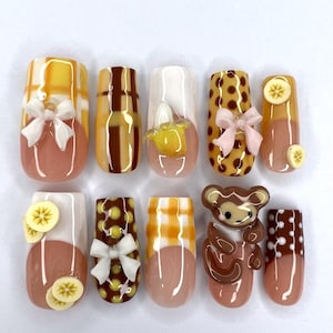 May include: A set of ten press-on nails featuring a variety of designs including yellow and brown plaid patterns, brown polka dots, white bows, miniature banana slices, and a three-dimensional brown monkey figure on a glossy, nude-coloured base.