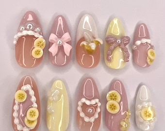Puede incluir: Un juego de diez uñas postizas en forma de almendra con acabado brillante en tonos rosa empolvado, amarillo pálido y blanco. Los diseños incluyen detalles tridimensionales como rodajas de plátano en miniatura, lazos blancos y cuentas de perlas.