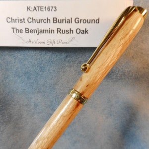 May include: A wooden pen with gold accents. The pen is made from a light brown wood with a natural grain pattern. The pen has a gold clip and a gold band around the barrel. The pen is labeled "K;ATE1673 Christ Church Burial Ground The Benjamin Rush Oak Heirloom Gift Pens".