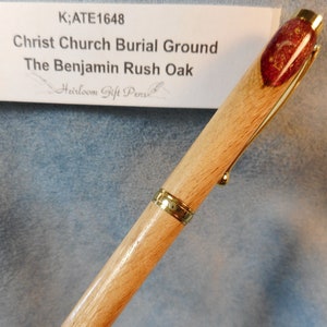 May include: A wooden pen with gold-coloured accents. The pen has a light-coloured wood barrel, a gold clip, and a decorative end with red and gold flecks. The text "Christ Church Burial Ground The Benjamin Rush Oak" is visible.