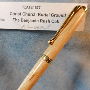 May include: A wooden pen with gold accents. The pen is engraved with the text "K;ATE1677 Christ Church Burial Ground The Benjamin Rush Oak Heirloom Gift Pens".