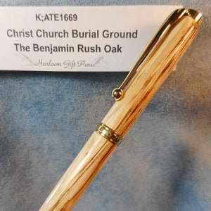 May include: A wooden pen with a gold-colored clip and accents. The pen is made from oak wood and has a natural wood grain. The pen is labeled "K;ATE1669 Christ Church Burial Ground The Benjamin Rush Oak Heirloom Gift Pens".