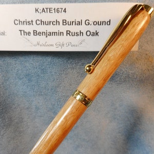 May include: A wooden pen with gold-colored accents. The pen has a light brown barrel and a gold clip. The text on the label reads "Christ Church Burial Ground" and "The Benjamin Rush Oak."
