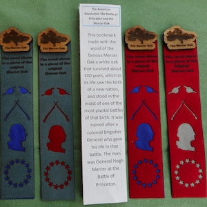 May include: Four wooden bookmarks with a red, white, and blue design. The bookmarks are made from the wood of the Mercer Oak, a white oak that survived for 300 years. The bookmarks feature a silhouette of a person, crossed rifles, and a circle of stars. The text on the bookmarks reads "The Mercer Oak" and "The wood above is a place of the original Mercer Oak".