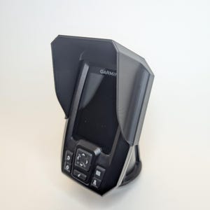 Może przedstawiać: Czarny echosonda Garmin z osłoną ochronną. Urządzenie posiada ekran, przyciski i podstawę. Słowo "GARMIN" jest widoczne na ekranie. Urządzenie jest przeznaczone do użytku w środowisku morskim.