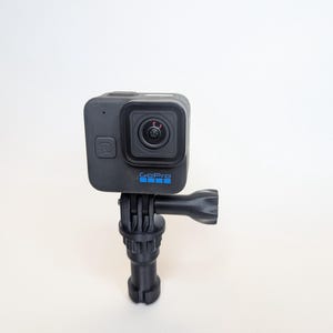 Puede incluir: Una cámara GoPro negra con el logotipo de GoPro en azul está montada en un trípode negro. La cámara tiene forma cuadrada con una lente en la parte delantera. El trípode tiene una rótula y una base para mayor estabilidad.