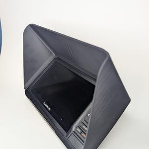 May include: A black Garmin device with a protective hood. The device has a screen and buttons labelled "BACK", "SELECT", and "MENU". The Garmin logo is visible on the screen. The hood is angled to provide shade.