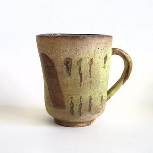 May include: A ceramic mug with a light brown glaze and a speckled texture. The mug has a rounded body and a handle. The glaze has a light green hue and brown streaks.