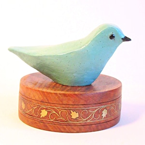 May include: A turquoise ceramic bird figurine with a black beak sits atop a round, wooden box. The box is reddish-brown with a decorative gold-colored floral band around its circumference. The bird has a speckled texture.