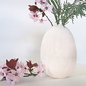 May include: White ceramic vase with a textured surface, shaped like an egg. The vase is filled with pink flowers and green foliage.