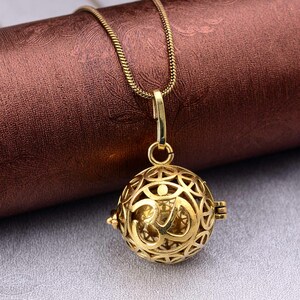 Op de afbeelding: Een goudkleurige hangerketting met een bolvormig, opengewerkt ontwerp. De hanger heeft een ingewikkeld patroon met een Om-symbool in het midden. De ketting heeft een fijne, goudkleurige schakel.