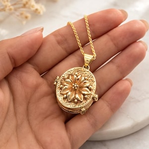 Könnte beinhalten: Eine goldfarbene Medaillon-Halskette mit einem ovalen Anhänger mit floralem Design. Der Anhänger hängt an einer filigranen Kette. Das Medaillon wird in einer Hand gehalten.