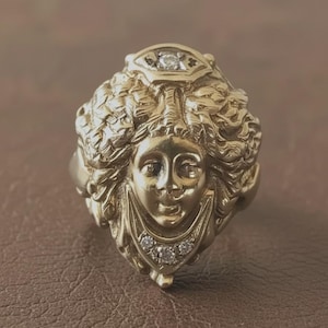 May include: A gold ring with a sculpted face design, showcasing intricate details in the hair and facial features. The ring is embellished with small, clear gemstones. The overall design suggests a vintage or antique style.