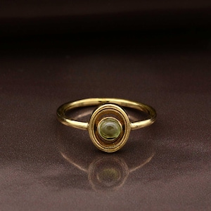Op de afbeelding: Een goudkleurige ring met een ovale zetting met een lichtgroene edelsteen. De zetting is omlijst door een cirkelvormig ontwerp. De ringband is slank en glad. Eenvoudig en elegant ontwerp.