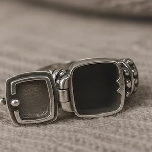 Könnte beinhalten: Ein silberfarbener Medaillonring mit einem offenen quadratischen Fach. Der Ring hat einen Klappdeckel, der sich öffnet, um einen kleinen Raum freizugeben. Das Band hat ein strukturiertes Design. Der Ring befindet sich auf einem neutralen Hintergrund.
