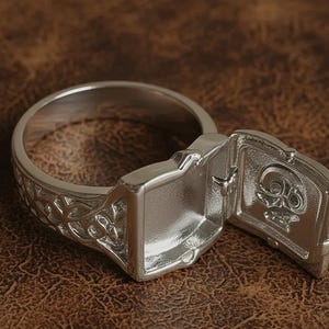 Op de afbeelding: Een zilveren ring met een open rechthoekig compartiment met een schedelontwerp. De ringband heeft een sierlijk bloemmotief. De ring wordt gepresenteerd op een bruin, leerachtig oppervlak.