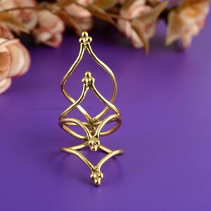 Puede incluir: Un anillo dorado para varios dedos con un diseño intrincado y calado. El anillo presenta una serie de bucles interconectados y elementos decorativos, creando una apariencia única y artística. El anillo está sobre un fondo púrpura.