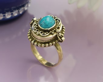 May include: A silver ring featuring a turquoise gemstone in an elaborate setting. The ring has a small, hinged compartment, and the band is plain, polished metal. The ring is shown on a purple surface.