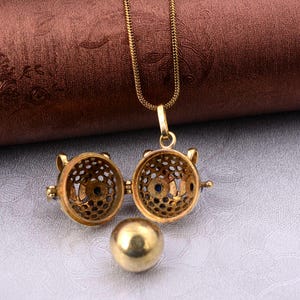 May include: A gold-colored pendant necklace with a detailed, openwork design. The pendant opens to reveal a small, blue gem. A separate gold-colored ball sits below the pendant. The necklace chain is also gold.