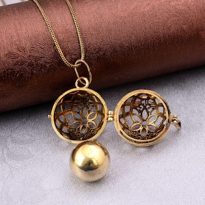 Puede incluir: Un collar con colgante dorado con un diseño de filigrana. El colgante se abre para revelar una pequeña esfera dorada. La cadena del collar es delicada y el colgante es redondo con un intrincado diseño calado.
