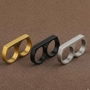 Könnte beinhalten: Drei Doppel-Finger-Ringe in Gold, Schwarz und Silber. Jeder Ring hat zwei kreisförmige Öffnungen für die Finger und einen rechteckigen Steg, der sie verbindet. Die Ringe werden auf einer braunen, strukturierten Oberfläche präsentiert.