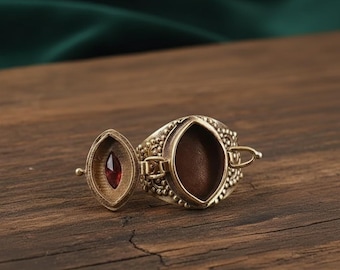 May include: A detailed, antique-style ring with an oval-shaped compartment. The ring is gold-toned with intricate detailing and a small, red gemstone. The compartment is open, revealing a dark interior. The ring is on a wooden surface.