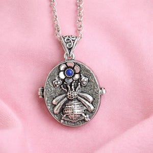 Puede incluir: Collar con colgante ovalado plateado con diseño de abeja y flor. El relicario presenta una abeja detallada con alas extendidas y una flor con una piedra azul en el centro. El collar tiene una cadena delicada y una fianza decorativa.