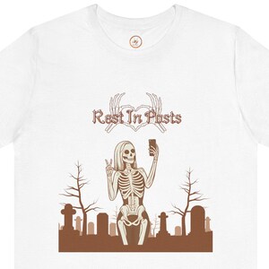 以下が含まれることがあります： 白のTシャツ。茶色のグラフィックで、墓地で自撮りをする骸骨が描かれています。骸骨は携帯電話を持ち、ピースサインをしています。「Rest In Posts」の文字が骸骨の上にあります。茶色の木と墓石が背景にあります。