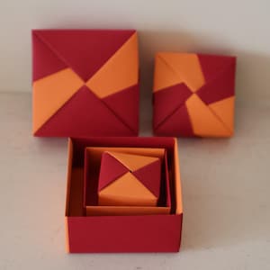 May include: A set of three origami boxes in red and orange. The boxes are square, with folded lids forming a geometric design. The largest box is open, displaying smaller nested boxes. The design showcases a striking contrast of colours and shapes.