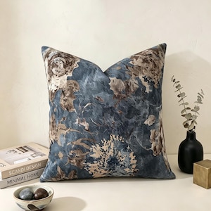 Può includere: Un cuscino quadrato in tessuto blu testurizzato con un motivo floreale astratto nei toni del marrone e del beige. Il cuscino presenta una cucitura visibile lungo i bordi e un aspetto morbido e imbottito, con un design intrecciato dettagliato.