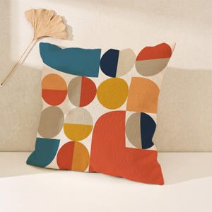 May include: A square throw pillow featuring a geometric pattern of circles, semi-circles, and rectangles in shades of teal, navy blue, mustard yellow, burnt orange, and beige. The design is printed on a textured, off-white fabric base.