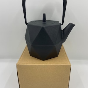 Puede incluir: Una tetera de hierro fundido negro con un diseño geométrico. La tetera tiene un asa y un pico. Está sobre una caja de cartón marrón. La tetera es de color negro mate.