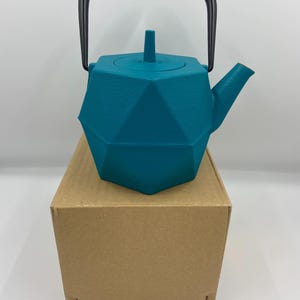 Puede incluir: Tetera geométrica turquesa con asa y boquilla negras. La tetera tiene tapa y está sobre una caja de cartón marrón. El diseño de la tetera presenta múltiples superficies planas, creando una apariencia facetada.