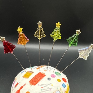 May include: A round pincushion with a fabric cover featuring sewing-themed patterns. Several decorative Christmas tree-shaped pins in red, orange, yellow, green, and clear are inserted into the cushion. Each tree has a star on top.