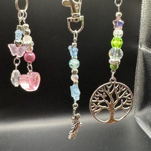 Op de afbeelding: Drie sleutelhangers met kralen en zilverkleurige sluitingen. Eén heeft een roze hartbedel en vlinderkralen. Een andere heeft een Vrijheidsbeeld-bedel. De derde heeft een levensboom-bedel en kleurrijke kralen.