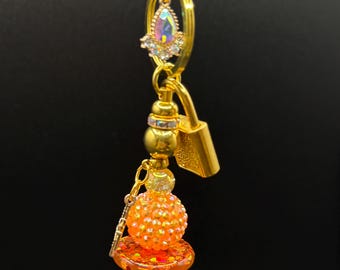 May include: A gold-colored charm with a teardrop-shaped iridescent gem and a small gold padlock. The charm features a series of beads in orange, green, and gold, with a small key dangling from the orange bead.