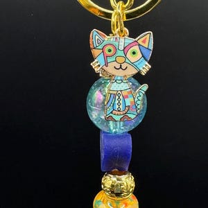 Puede incluir: Un llavero con forma de gato colorido con un anillo dorado. El colgante de gato presenta un diseño geométrico en varios colores. El llavero incluye un orbe azul, un rectángulo morado y otras cuentas decorativas.