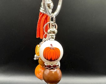 May include: A fall-themed keychain with a silver ring and orange tassel. The keychain features a white bead with an orange pumpkin design, brown and orange beads, and silver accents. A festive accessory.