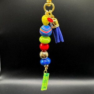 May include: A colorful beaded keychain with a gold clasp and accents. The beads are yellow, blue, green, red, and blue, with a small blue tassel. A neon green rectangular charm hangs at the bottom.