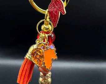 May include: A gold-toned keychain with autumn-themed charms. It features a red leaf charm, an orange leaf charm, and a tassel. The keychain also includes a variety of beads in gold, black, and orange.