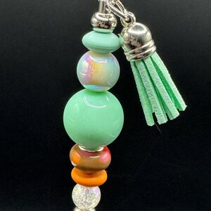 以下が含まれることがあります： シルバーチェーンとミントグリーンのタッセルが付いたビーズキーホルダー。キーホルダーは、緑、虹色、オレンジ、透明なビーズなど、カラフルなビーズのスタックが特徴です。ビーズはさまざまな形とサイズです。