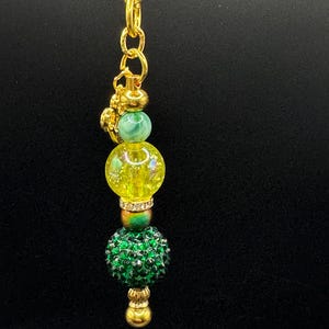 Puede incluir: Un llavero dorado con un cierre de mosquetón. El llavero presenta una serie de cuentas decorativas en verde y amarillo, con detalles dorados. Las cuentas incluyen una cuenta verde veteada, una cuenta amarilla con purpurina incrustada y una cuenta verde de pedrería.