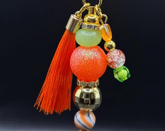 May include: A gold-colored keychain with a decorative charm. The charm features an orange tassel, orange glittery bead, green bead, and various other colorful beads. The keychain is designed with a gold ring and a black background.