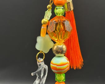 May include: An orange beaded charm with a clear high-heeled shoe pendant. The charm features orange, gold, green, and clear beads, a small orange tassel, and a gold-colored clasp. The background is black.